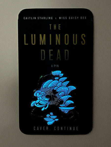 A Luminous Dead horror skull rainbow metal enamel pin with glowing blue mushrooms growing out of its orifices. The backing card is of a blue cave, and it says Caitlin Starling x Miss Daisy Dee, The Luminous Dead, a Pin, "Caver, continue." On a darkened white background.