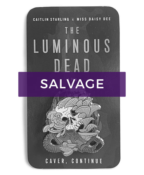 Image fully desaturated, with a purple "Salvage" label overlaid. A Luminous Dead horror skull pin with pale teal blue mushrooms growing out of its orifices. The backing card is of a blue cave, and it says Caitlin Starling x Miss Daisy Dee, The Luminous Dead, a Pin, "Caver, continue." On a white background.