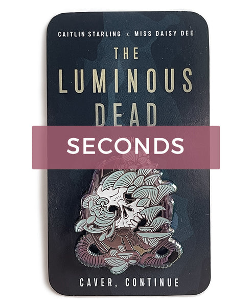 Image desaturated and overlaid with a "Seconds" label. A Luminous Dead horror skull pin with pale teal blue mushrooms growing out of its orifices. The backing card is of a blue cave, and it says Caitlin Starling x Miss Daisy Dee, The Luminous Dead, a Pin, "Caver, continue." On a white background.