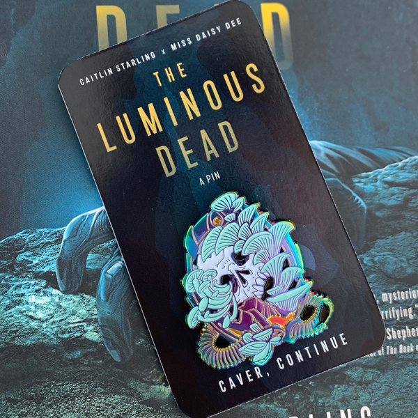 A Luminous Dead horror skull pin with pale teal blue mushrooms growing out of its orifices. The backing card is of a blue cave, and it says Caitlin Starling x Miss Daisy Dee, The Luminous Dead, a Pin, "Caver, continue." With the original novel cover as the background.