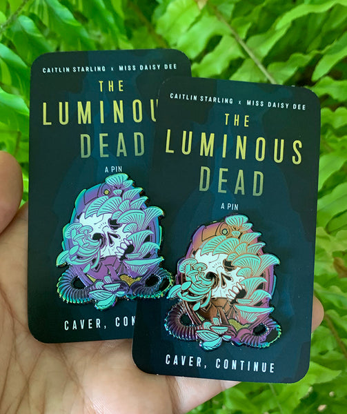 Image of two rainbow skull pins on their backing cards in front of a fern, being held up by a hand. they are displaying the variety in metal colors and the left one is mostly blues and purples in the metal, while the riht one has purples, pinks, and orange metal colors as well. 